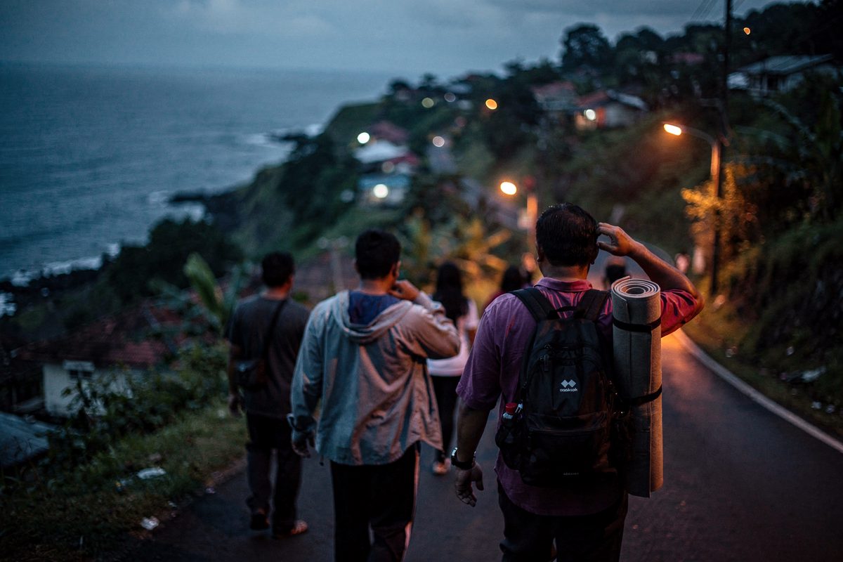 Pre-dawn on a hillside road above a dark Indonesian coastline, figures seen from behind walking uphill away from the water, one person carrying a rolled.