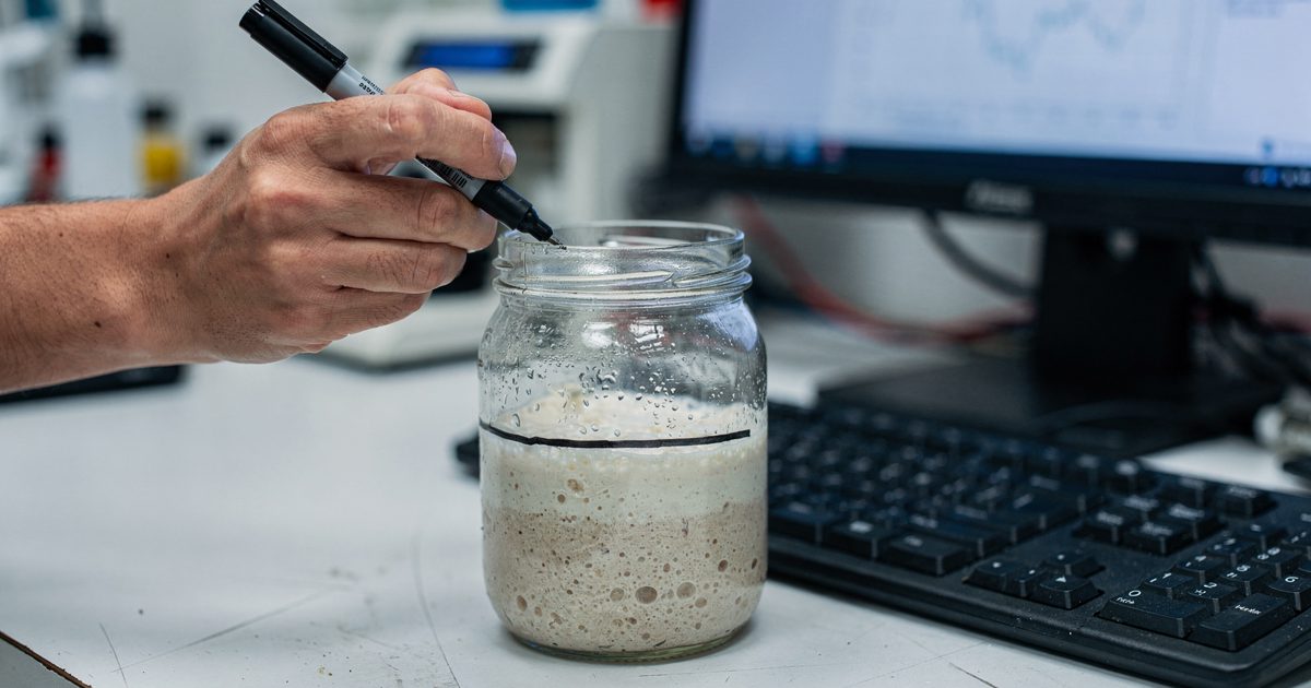 Close detail under bright fluorescent laboratory light at mid-morning, a technician's hand cropped at the wrist drawing a fresh pen line on the side of a glass.