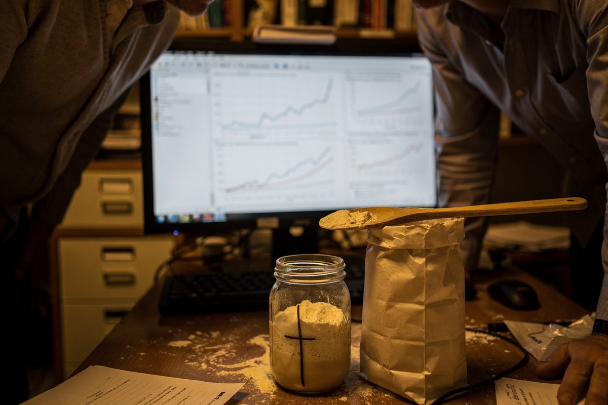 Medium shot in late evening amber light inside a cramped university research office, two researchers seen only from the shoulders down leaning over a desk.