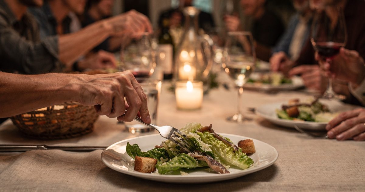 Table-height shot by candlelight at a crowded home dinner party, a guest seen only from the forearm down setting a fork beside a half-eaten caesar salad while.