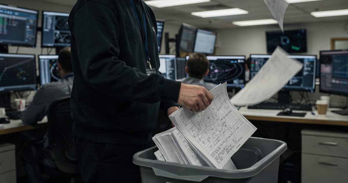 Under harsh fluorescent lighting in a windowless mission control center, a technician shot from behind drops a thick stack of trajectory printouts directly.