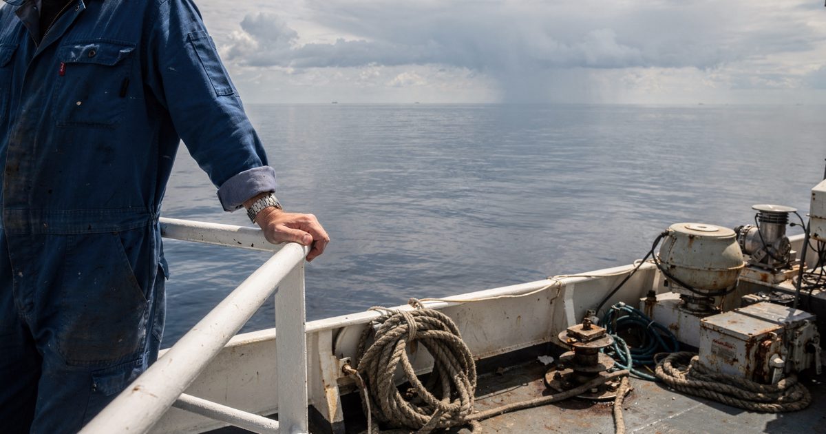 Medium shot in harsh midday glare on the deck of a research vessel, a scientist seen from the chest down in a blue boiler suit leaning against the guardrail to.
