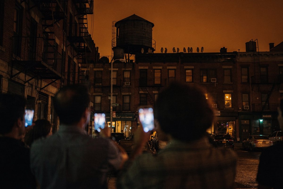 Low-angle shot from street level at night on an Upper West Side sidewalk, a small crowd seen from behind with heads tilted back and phone screens raised toward.