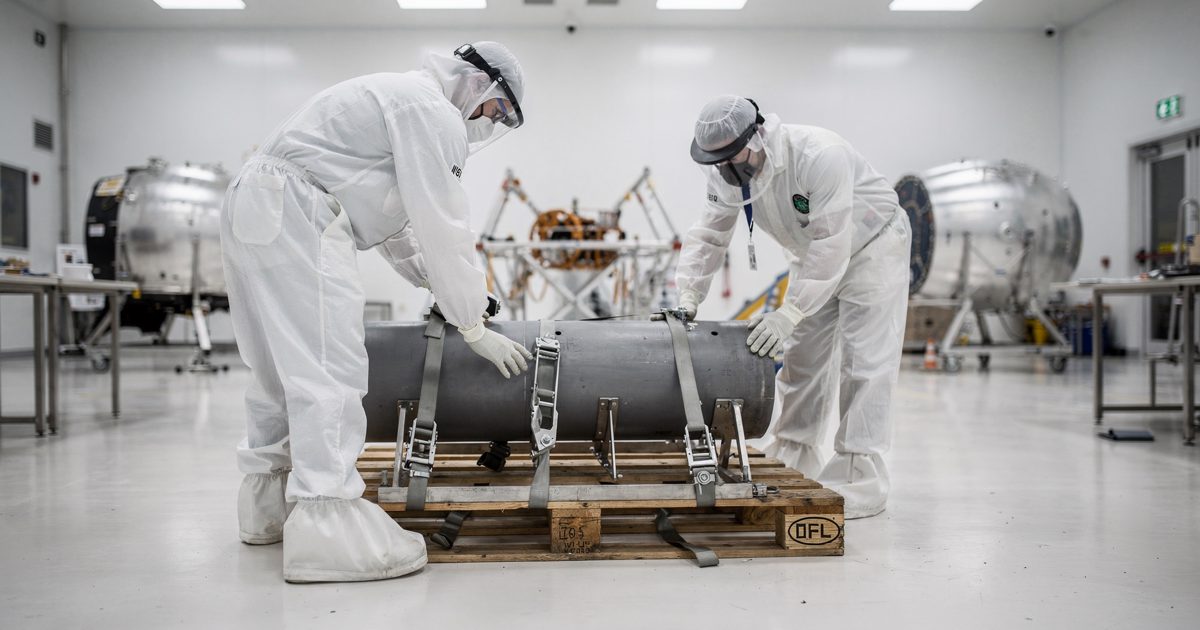 Wide shot inside a sterile white aerospace clean-room under bright shadowless LED panels, two engineers in full protective suits seen from behind strapping a.