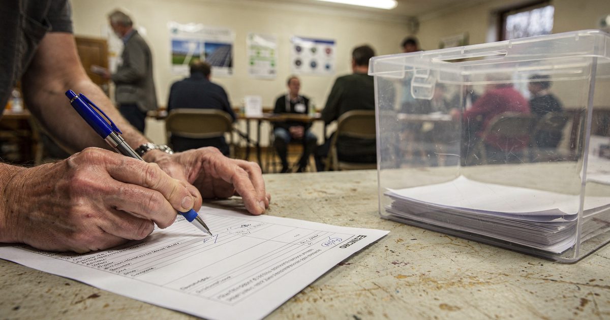 Close detail under bright fluorescent light in a village hall drop-in session, a resident's hands cropped at the wrists pressing a ballpoint pen into a paper.