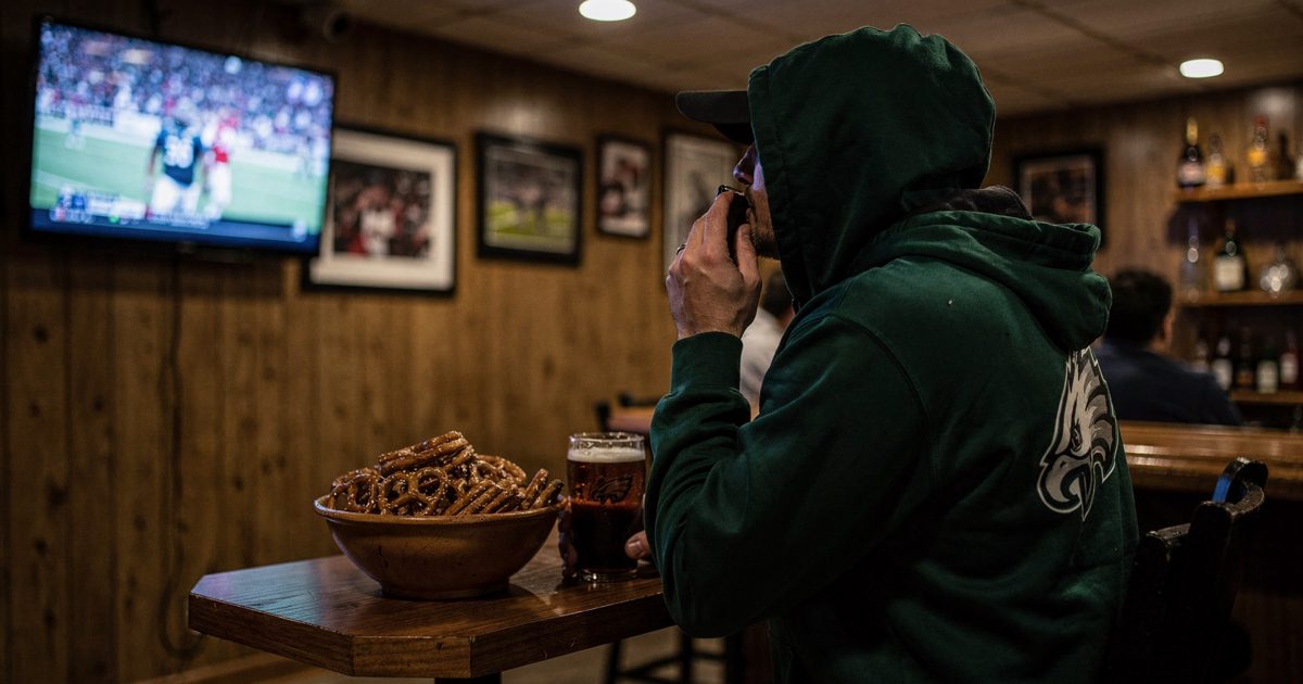 Eye-level shot in a wood-panelled suburban basement bar late at night, a man in a green Eagles hoodie with his face turned toward a glowing wall-mounted TV.