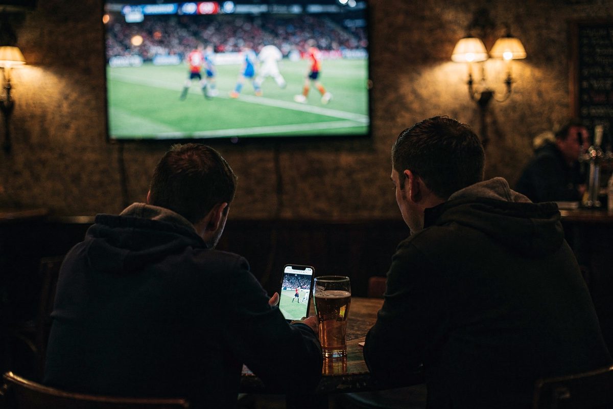Dim pub interior in Holloway at night with a wall-mounted television showing a muted football match, three patrons seen from behind crowding around a single.