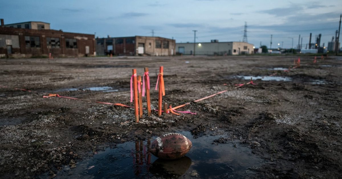 A wide dusk photograph of a vacant industrial lot beside low warehouses, orange survey stakes in wet earth, a scuffed football half-submerged in a puddle in.