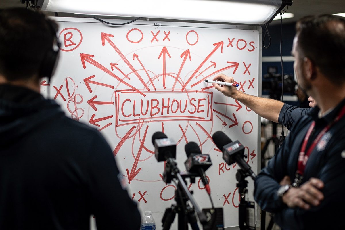 Close detail inside an NFL media room under harsh LED spotlights, a dry-erase board scrawled with aggressive red defensive coverage arrows converging on a.