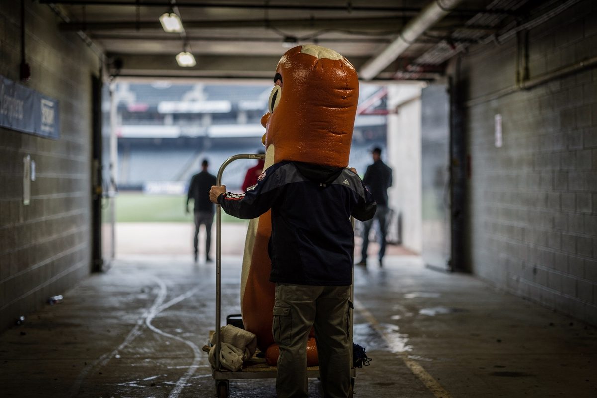 Medium shot from behind in a concrete ballpark service tunnel under caged bulbs, a handler seen from the shoulders down steadying a towering foam sausage.