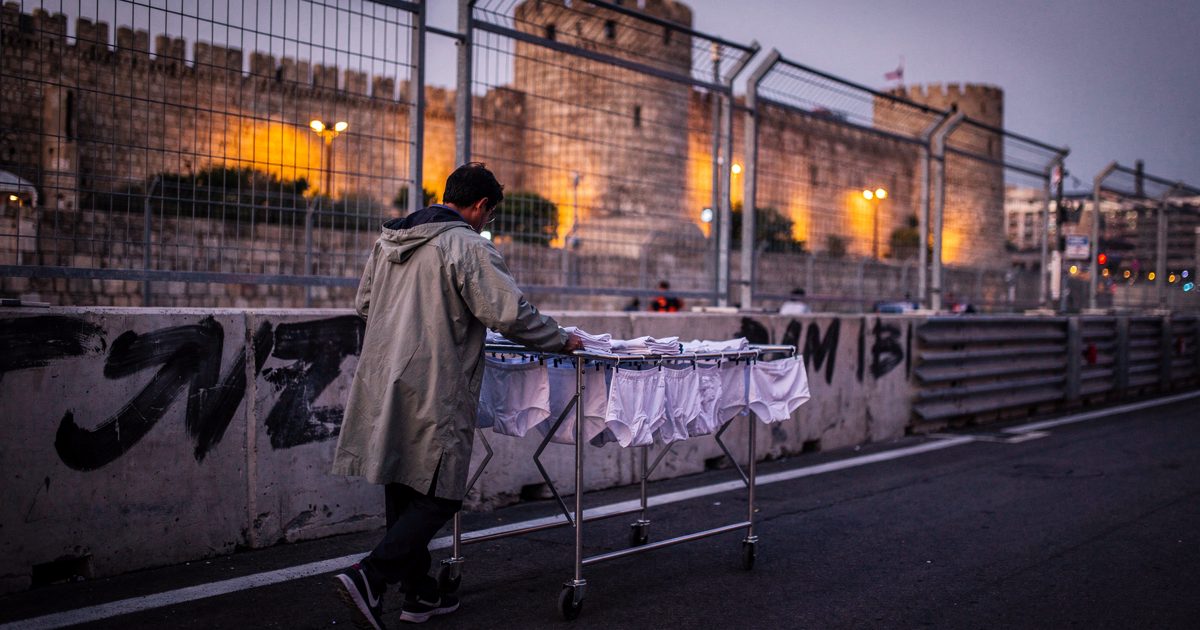 Wide shot at violet dusk along the Baku street circuit's concrete barriers below the old city walls, a figure seen from behind in a raincoat pushing a wheeled.