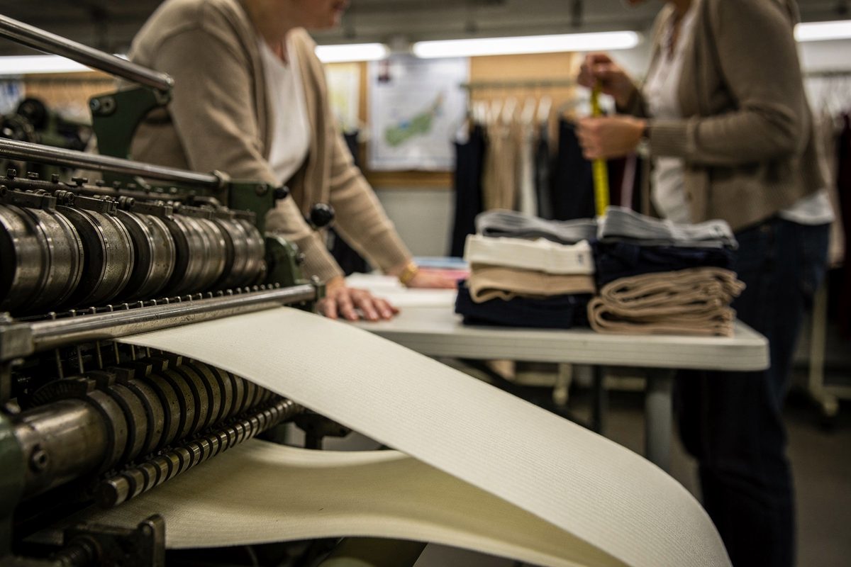 Low-angle mid-shot under humming fluorescent strip lights on an East Midlands hosiery mill floor, the torsos of several people in cardigans blurred around a.