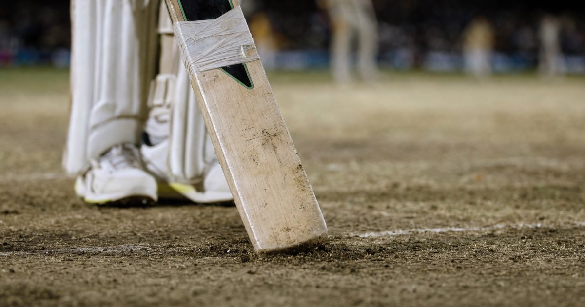 A close-up ground-level shot of a scuffed, dusty cricket pitch under harsh stadium floodlights, with the lower half of a battered, tape-wrapped wooden cricket.