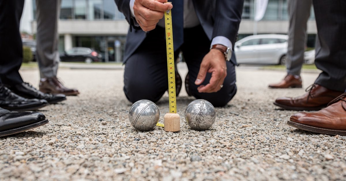 Close low-angle shot across pale gravel in flat midday light as a kneeling official stretches a measuring tape between two steel boules and a small wooden.