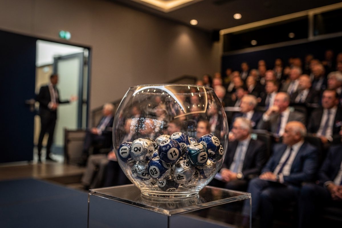 Interior shot under warm stage lighting of a glass bowl of numbered draw balls sitting untouched on its plinth, rows of seated delegates blurred and turned.