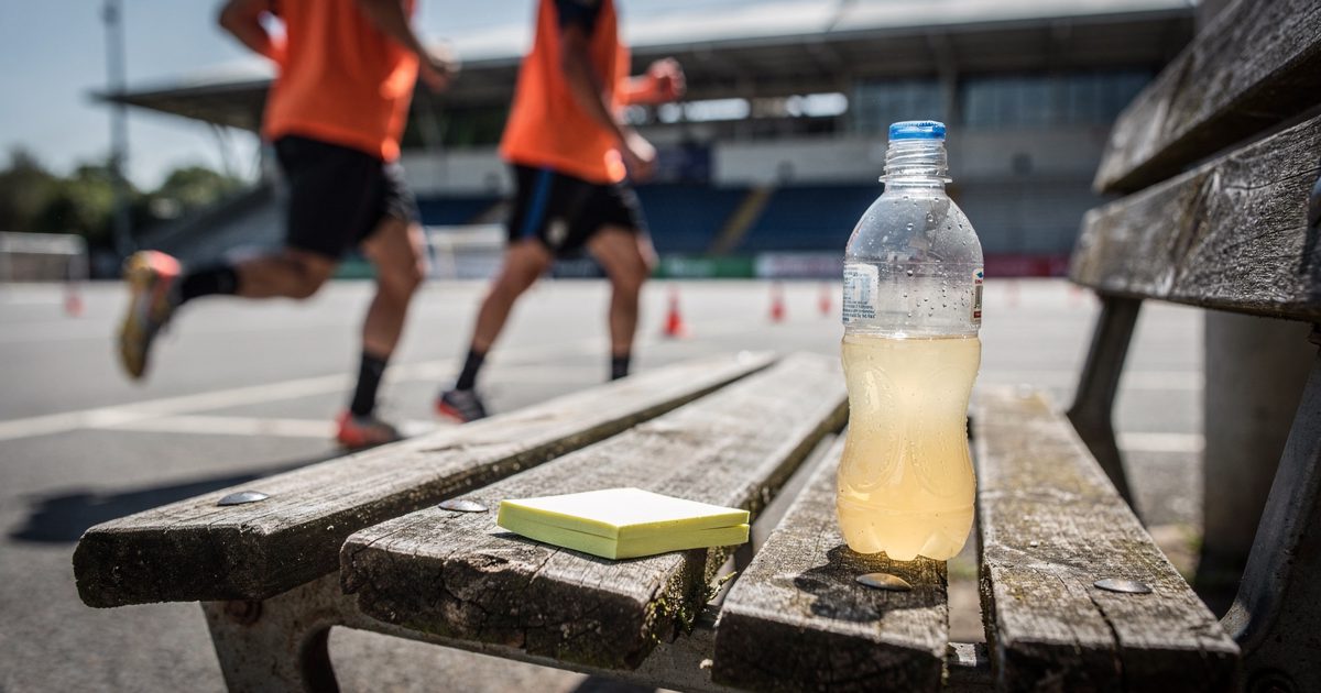 Close detail in harsh midday sun on a stadium car park bench, a yellow sticky note stuck to the slat beside a half-drunk sports drink bottle, its handwriting.