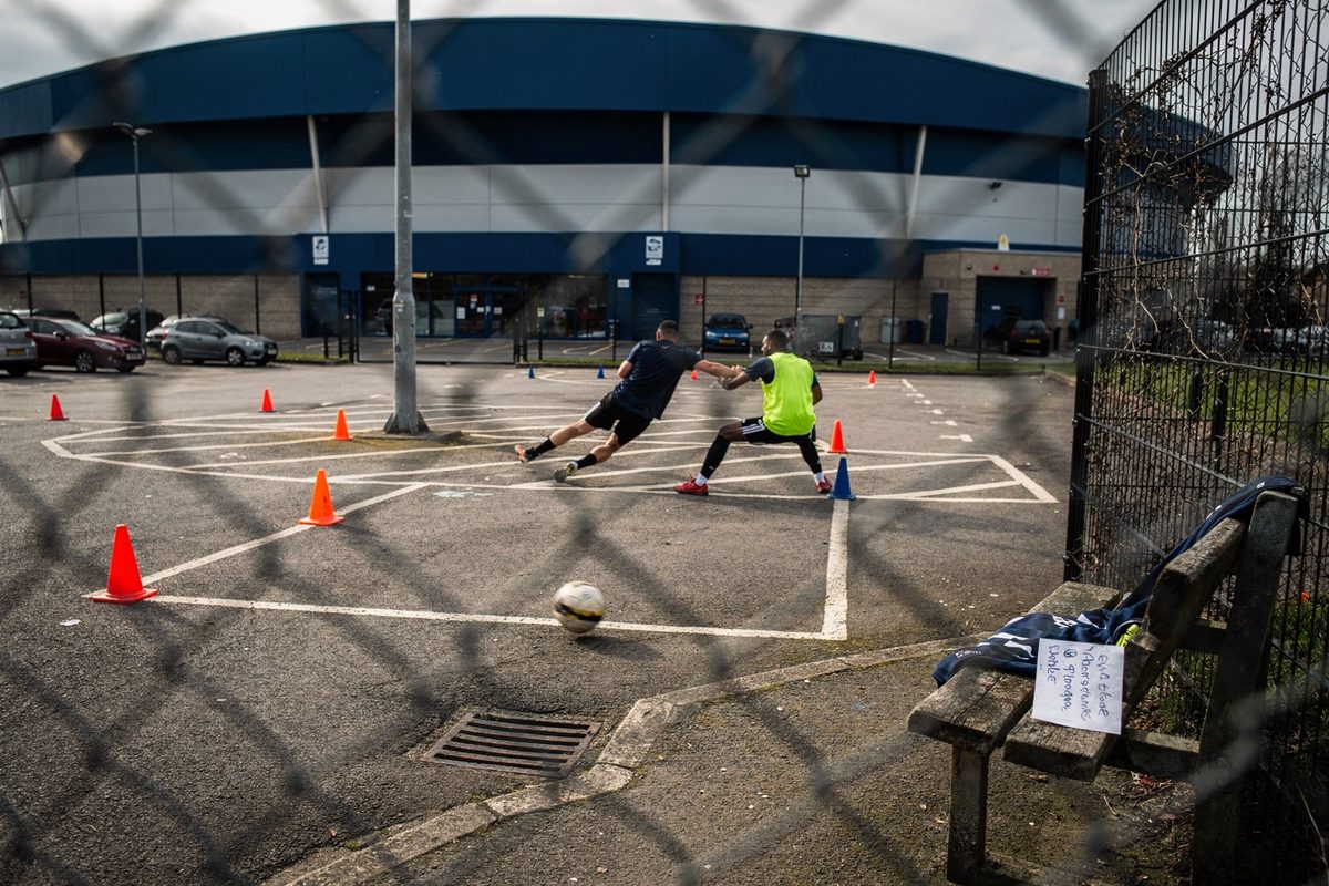 Wide shot through a chain-link fence at noon of a five-a-side pitch marked out with traffic cones between painted parking bays behind a football stadium.