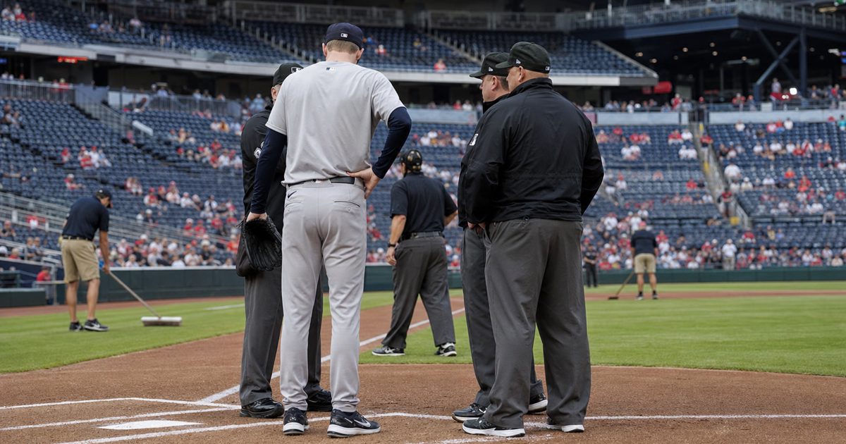 A realistic editorial sports photograph inside a major-league baseball stadium before a game, showing an unidentified uniformed pitcher from behind speaking.