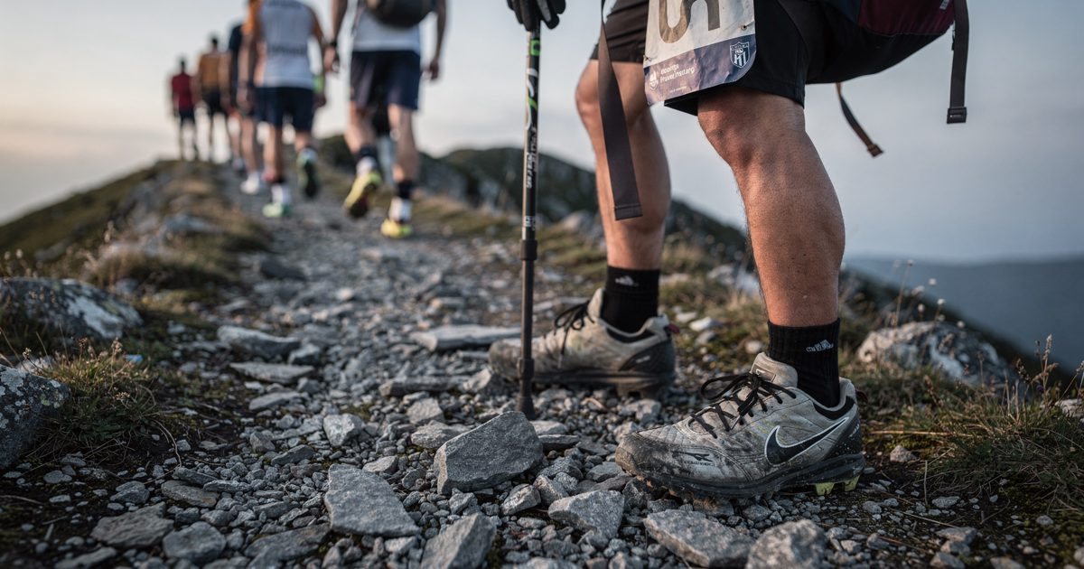 Close on a pair of football boots planted on loose scree in late side-light, one hand gripping a hiking pole while a numbered training bib flaps from a.