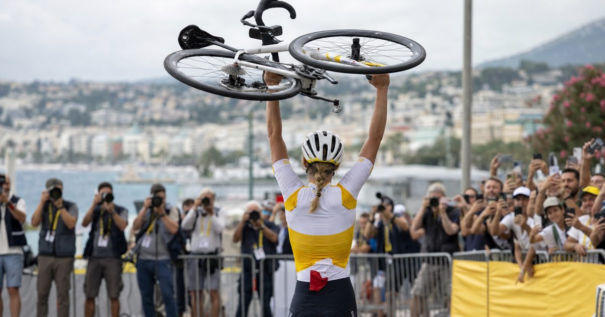 News photograph at a professional women’s cycling finish area in Nice, a victorious rider seen only from behind lifting a road bicycle overhead, crowd barriers.