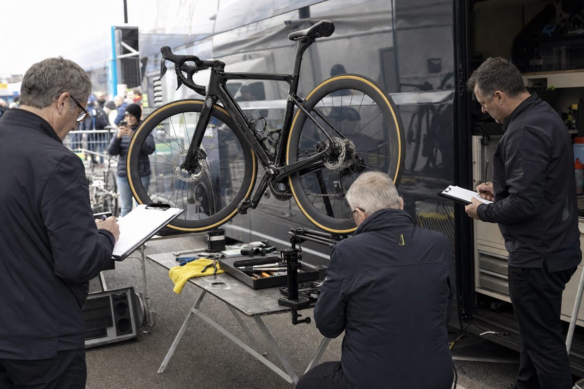 Documentary-style photograph of anonymous cycling technicians examining a high-end road bicycle on a folding inspection stand beside team vehicles, clipboards.