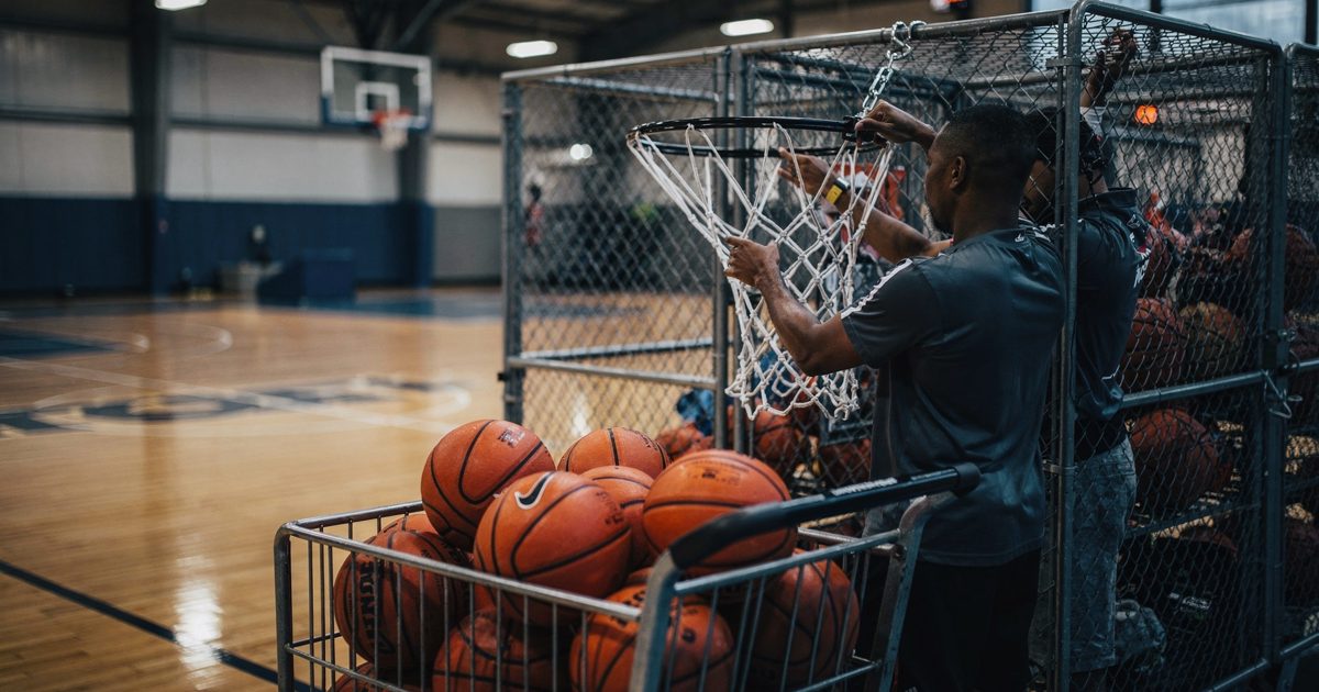 Early evening fluorescent banks over an indoor NBA practice court as two staffers cropped at the neck pull a spare white basketball net toward a hook on a.