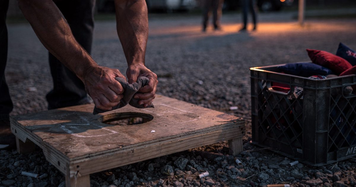 Close-up at dusk of two cracked hands gripping a bean bag above a plywood cornhole board on a gravel lot, low orange light from a single pole lamp, a milk.