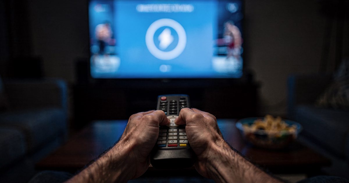 A viewer's hands, tense and white-knuckled, crush a plastic remote control in a darkened living room illuminated entirely by the stark blue glare of a.