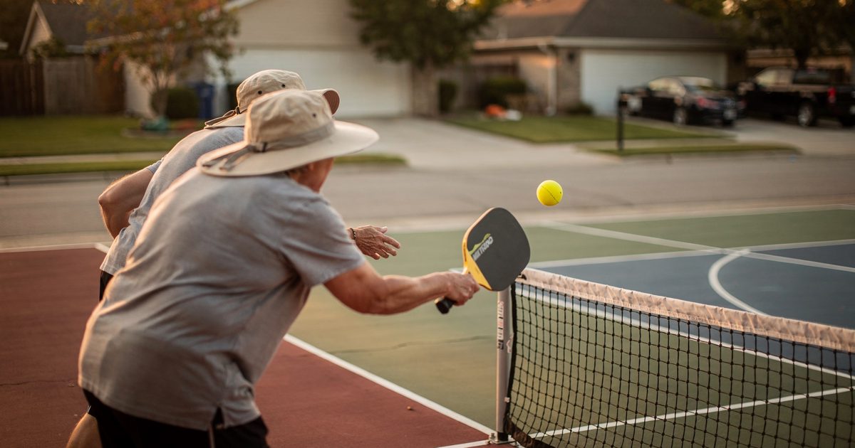 From court level in late-afternoon gold on a shared cul-de-sac pickleball court, two neighbors in sun hats lean through a serve, a split ball just leaving the.