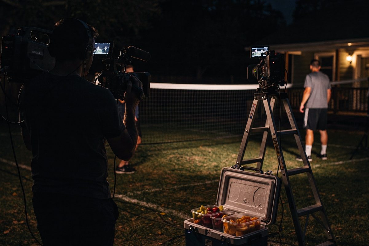 Over the shoulder of a camera operator at full dark, porch lights raking a net while two players hold opposite baselines and a folding ladder stands beside a.