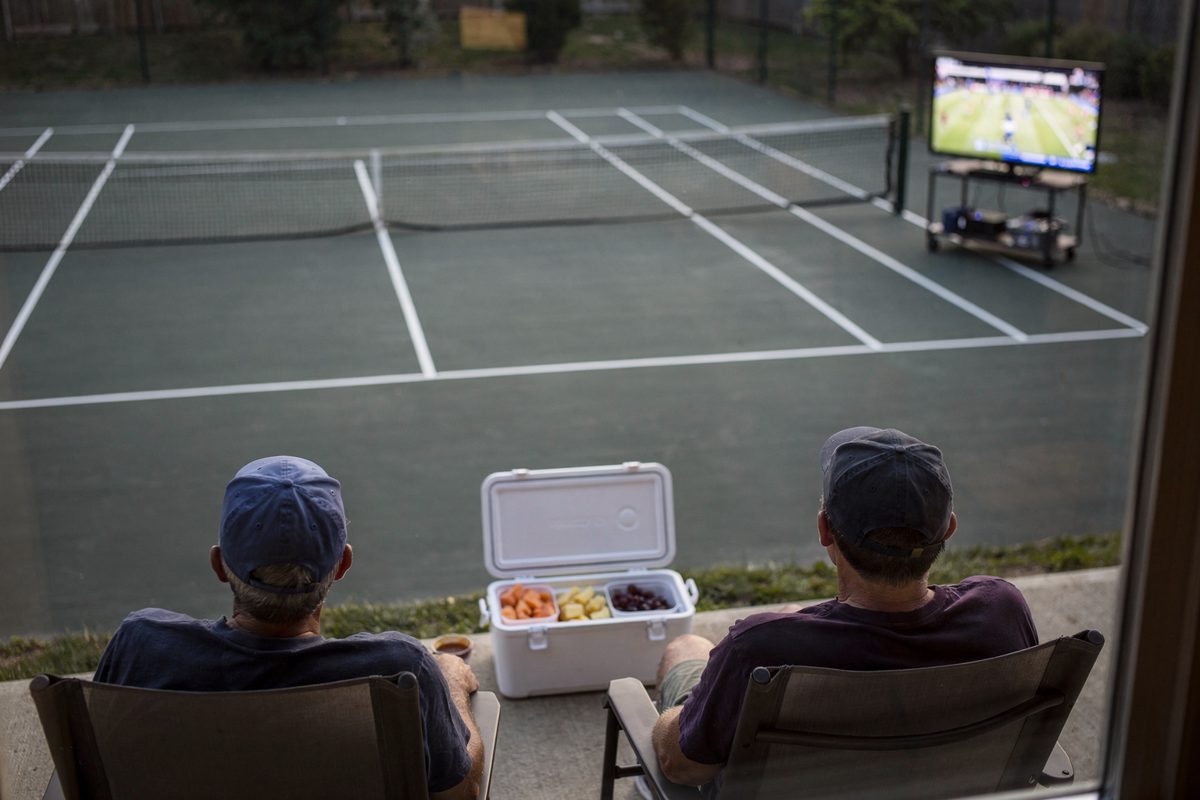 From a second-story window in pale morning light, three neighbors visible only as caps sit in folding chairs facing an empty court, an open cooler of cut fruit.