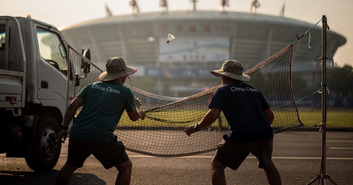 Medium shot from a low angle in hot late-afternoon sun on the service road behind a tennis stadium in Beijing, two grounds staff in work polos and sun hats.