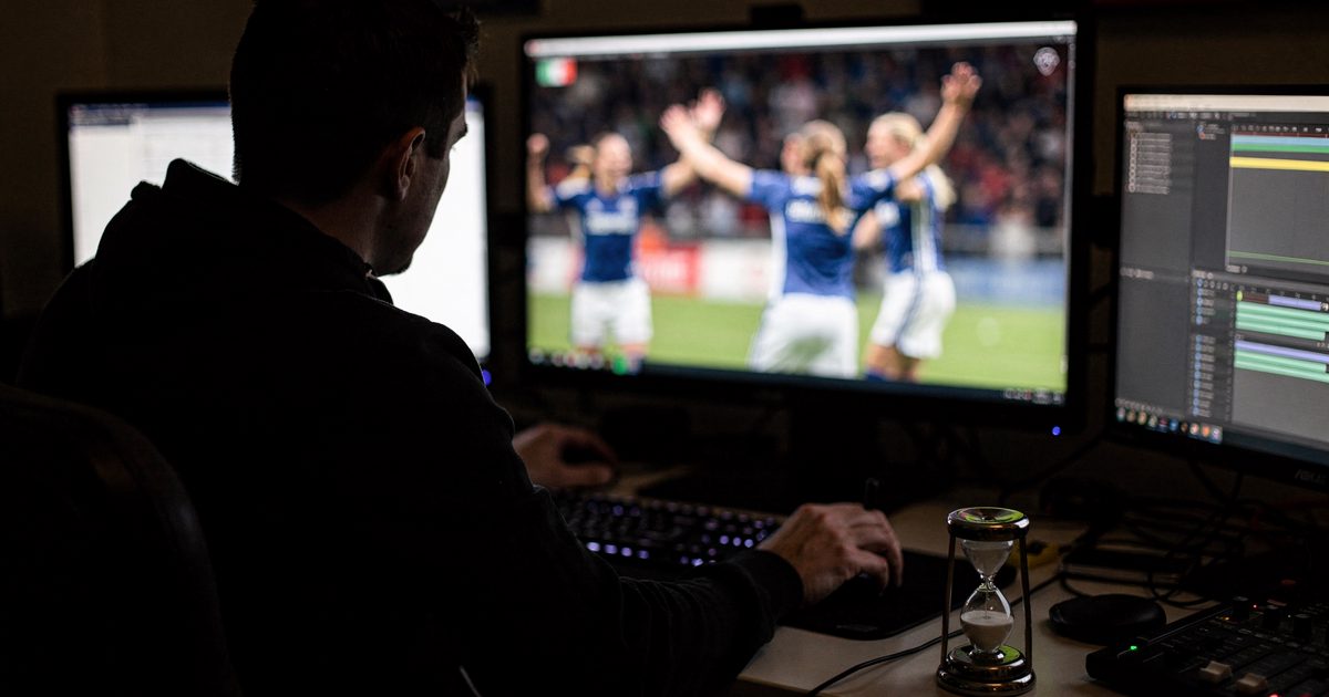 Medium shot inside a windowless broadcast edit suite lit only by monitor glow at midday, a video editor seen in silhouette against the screen leaning in to.