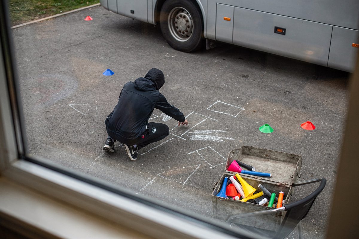 High angle through a canteen window in flat morning light, looking down on a tarmac bay where two tracksuit figures with hoods up and faces hidden chalk six.