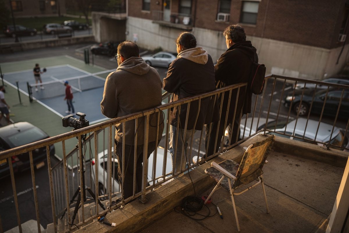 Warm late-afternoon light on a concrete stairwell landing above a parking lot, five residents seen from behind pressed against the metal railing watching a.