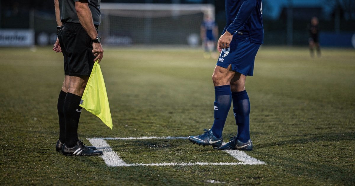 Dusk floodlights throw long shadows across a club training pitch as a match official cropped at the chest holds a yellow flag at waist height beside a white.