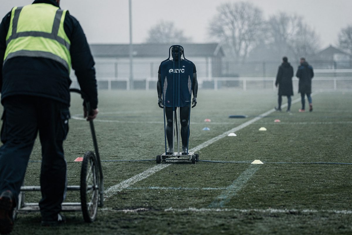 Pale morning light over a club training pitch as a grounds worker cropped at the shoulders wheels a single mannequin toward the centre-forward spot, two.
