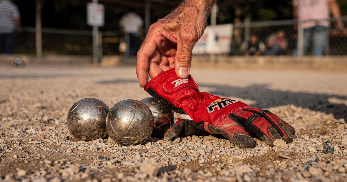 Close detail in hard late-afternoon Madrid sun on a crushed-gravel petanque court, a red racing glove lying beside three scuffed steel boules while a.