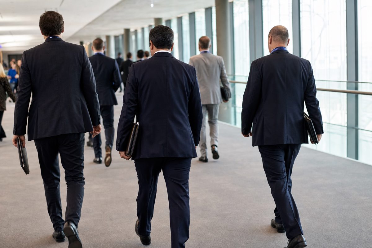 A documentary-style photograph of anonymous suited sports administrators walking briskly through a modern convention centre while carrying document wallets and.