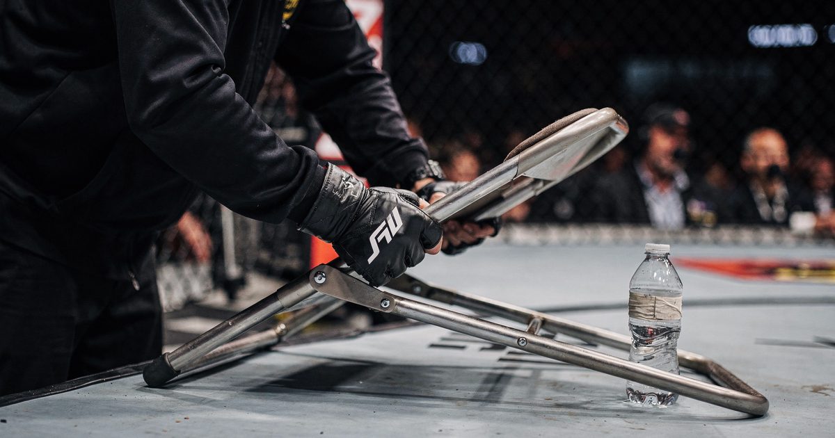 Cage-side under harsh pre-fight arena lights, a close crop of gloved hands and a part-cropped cornerman slamming a metal folding chair open on the canvas next.