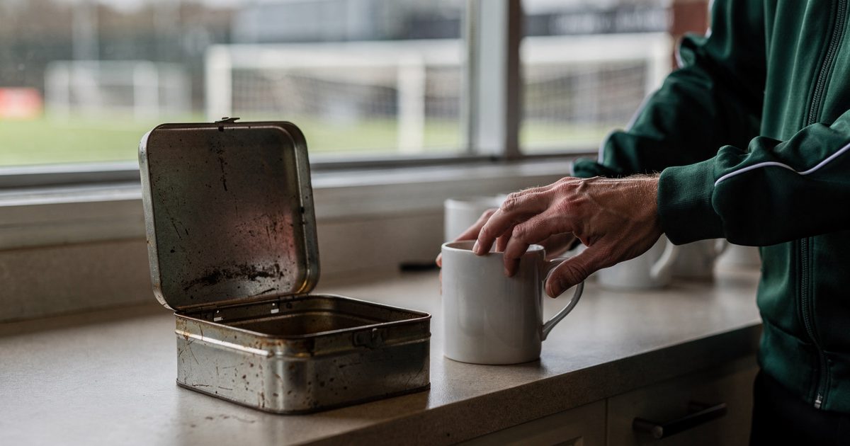 Close-up of a hand setting a white mug beside an open metal biscuit tin on a canteen counter at a football training ground, late morning light from high.