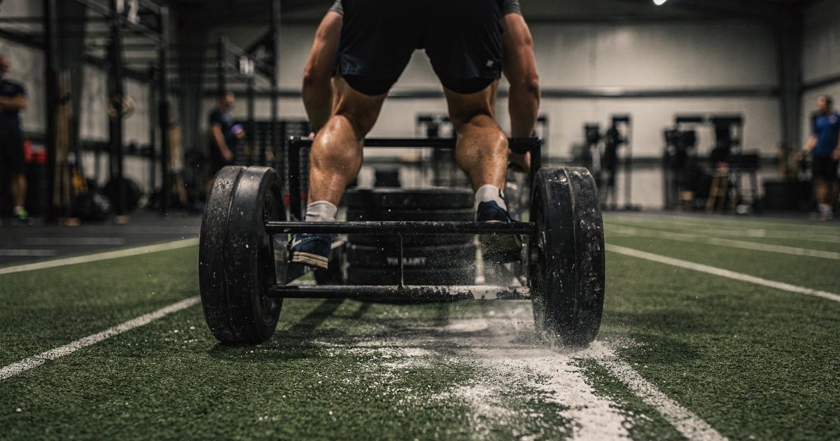 Low-angle shot in harsh gymnasium lighting of an athlete seen from behind pushing a heavy black sled across a scuffed indoor track, a trail of white chalk dust.