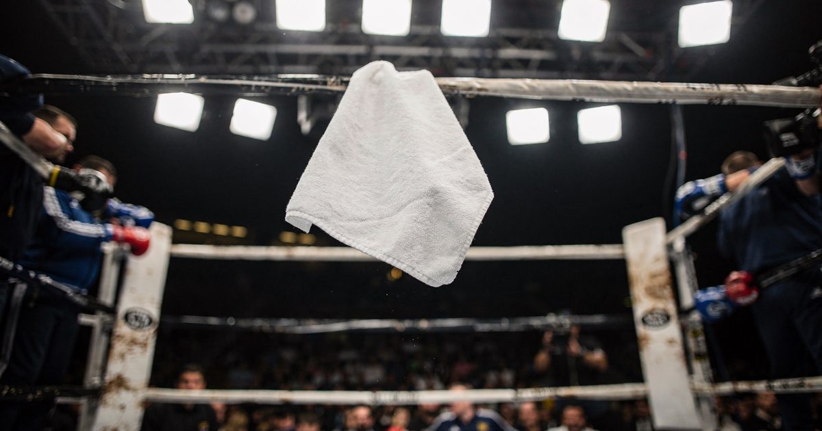 Night arena interior shot from below the ring apron, a white towel frozen mid-air above sagging ropes under television light rigs, cornermen visible only as.