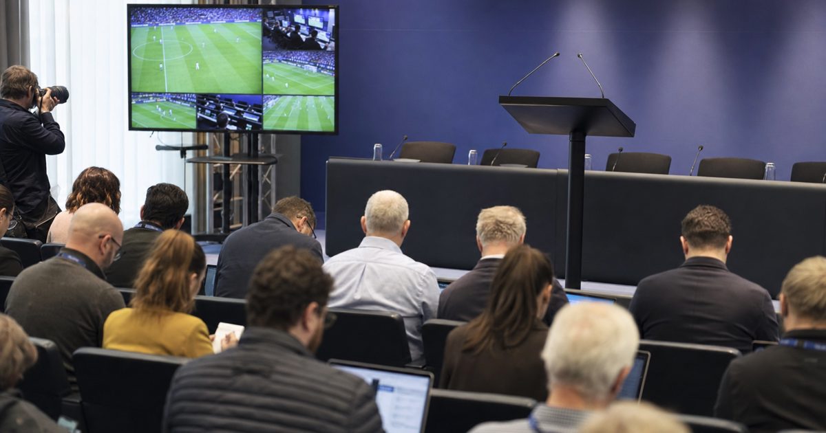A formal football governing body press room with an empty lectern, rows of journalists holding notebooks, and a large referee review monitor beside the stage.