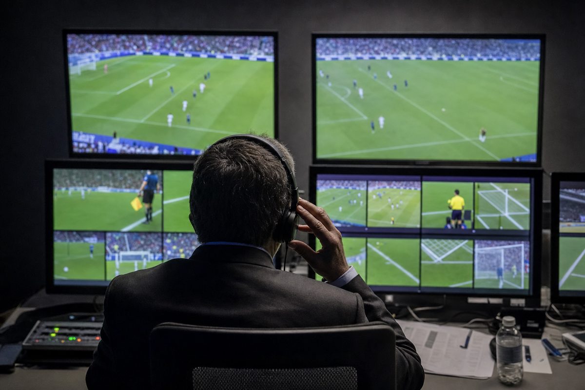 A lone suited official viewed from behind in a dim video review booth, studying several monitors while touching an earpiece, realistic sports news photography.