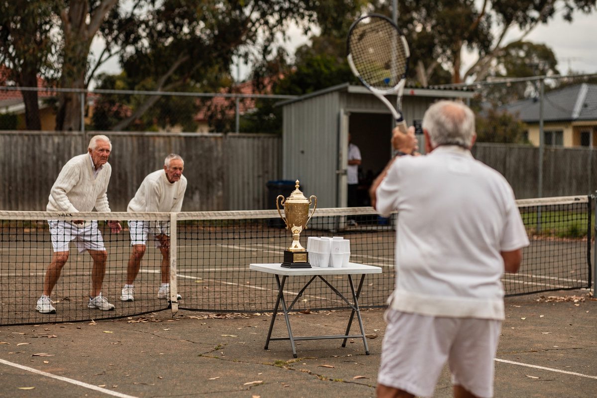 Wide shot of a modest Australian suburban tennis club court framed by gum trees, four part-cropped older players in white knitwear mid-rally over a sagging.
