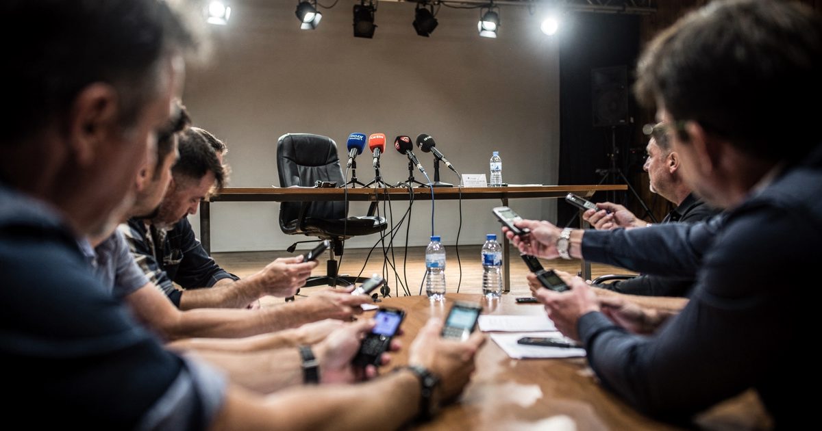 Low angle from the floor of a crowded Belgrade media center under harsh spotlights, a black office chair spinning empty behind a table covered in foam-tipped.