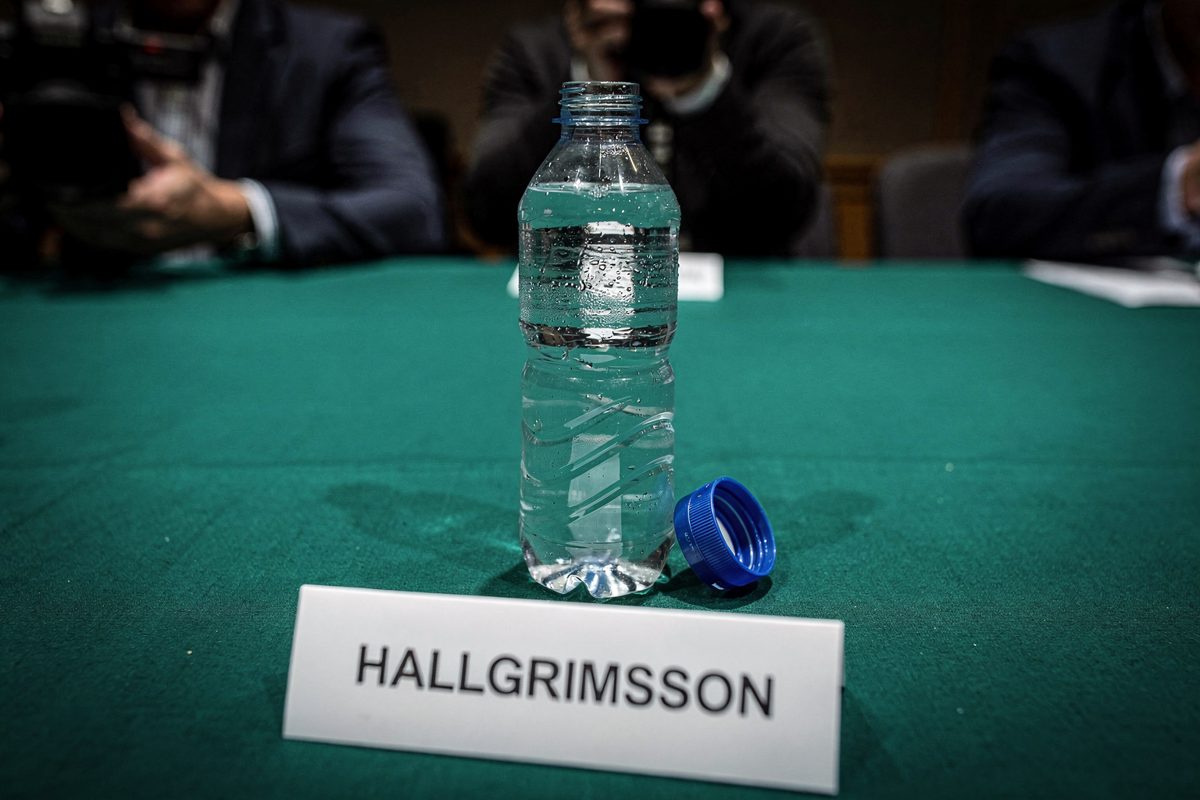 Close detail in stark overhead flash lighting on a green felt-covered table, a plastic water bottle sitting with its blue cap resting entirely unscrewed beside.