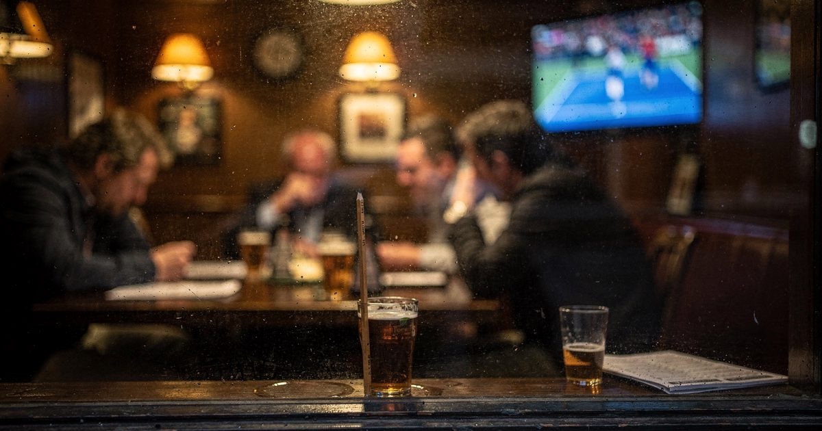 Medium shot from the street through a fogged pub window late on a dark evening, blurred figures leaning over a crowded table under amber wall lamps while a.