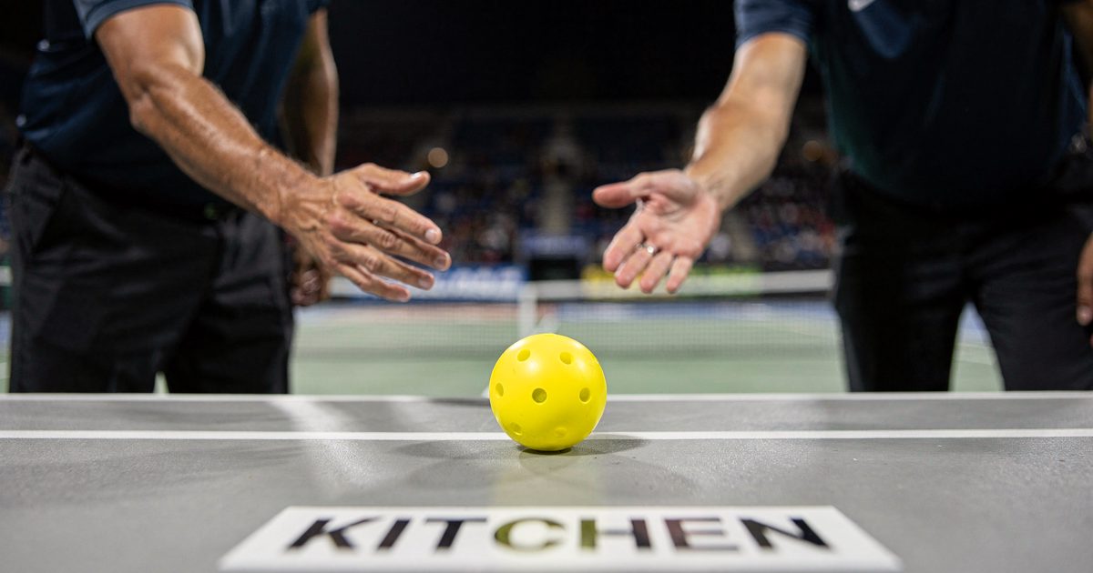 Close night shot under hard stadium floodlights of a yellow pickleball resting on a white kitchen line on a portable court, two officials' hands hovering above.